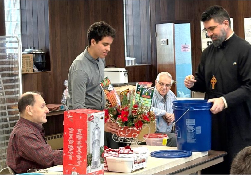 Fr. Aleksey is standing at the end of a table, with his left hand on a blue, five-gallon bucket, and he's reading a raffle ticket that's in his right hand. A young man is standing around the corner of the table, looking towards the bucket, and two men are seated on either side of him.