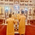Four clergy members, in two rows, stand behind Fr. Aleksey, who is just inside the Royal Doors at the altar. Their backs are facing the camera as they look towards the altar and iconostas. There is a gold cross and an icon on a table between the two rows of clergy, who are all wearing gold vestments.