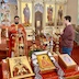 Father Aleksey is standing at the front of the church, facing the congregation. He's holding the communion chalice in his left hand, and a red cloth is draped over his right hand. A male communicant is waiting off to his left-hand-side, and a table with three icons, a cross, flowers, and candles appears in the photo's foreground.