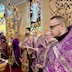 Close-up photo of six priests standing in a line along the back wall of the altar. The photo was taken from the right side of the altar, and a large cross appears in the upper right corner of the frame, with the altar table just slight visible in the lower left corner.