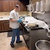 The man from the previous photo is now standing in front of an industrial-sized combination stove-oven, which appears in the right half of the frame. He's holding a metal bowl with a ball of dough in it, and there's another metal bowl with dough on the stovetop, and its covered with cellophane so the dough can rise. Kitchen cabinets are visible in the background of the left side of the photo.