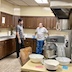 Father Aleksey is standing and talking to the man from the previous two photos. They're standing at the back of the kitchen, and the man is leaning against the countertop and lower cabinets. A table with a dough mixer and three white bowls are visible in the foreground, and there's a very large, brown bag (I'm guessing 25 lbs) of flour resting on a chair on the left side of the table.