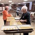 Two women are standing on opposite sides of a long, brown table that runs mostly perpendicular to the bottom edge of the frame. Using their gloved hands, they are rolling dough pieces into shapes that will be placed on top of the paska breads before baking. The woman on the left side is further away from the camera. She's wearing an orange sweatshirt and is making long rolls of dough that look kind of like snakes. The rolled lengths will then be braided and and placed around the circumference of the breads. The woman on the right is closer to the camera. She's wearing a long-sleeved, black shirt and is making shorter rolls of dough and putting them together to form Orthodox crosses. In the foreground there are two metal, cookie trays that are perpedicutlar to the bottom frame. The one on the left contains finished dough crosses, and the one on the right has finished dough braids. 