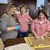 Two women and a teenage girl are standing at a brown table with a slicon mat in front of the girl. The woman on the left and the girl, who is standing on the right, are looking at the mat, while the woman in the center is smiling and looking at the camera. The woman on the left is wearing a grey shirt with 3/4-length sleeves and blue food-prep gloves. The girl is wearing a pinksih hoodie with the sleeves pushed up to her elbows, and she also has on clear food-prep gloves. The woman in the center is wearing dark blue jeans and a red and white striped shirt with skinny stripes. Her hands are not visible in the photo. Oh yeah, and the two women have sunglasses resting on the tops of their heads.