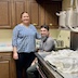 Matushka Natallia is standing next to Father Aleksey, who is seated in the back corner of the hall kitchen, between the far end of the stove and the back countertop and cabinets. She's wearing a blue and white oval-patterned shirt and black pants, and she has her left arm resting on Fr. Aleksey's right shoulder. He's wearing a gray shirt and gray pants. They're both smiling and looking at the camera. 