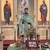 Father Aleksey is standing in front of the Royal Doors, facing the congregation. He's wearing a green vestmenet and is holding a white cross in his right hand. Visible in the foreground, is the center table, which has an icon, two vigil lights, a gold cross and gold bowl, a brown binder, and pussy willows on top of it. The icon is surrounded by a purple cloth that serves as a frame.
