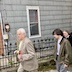 Part of the procession line outside of the church, where three people are visible, walking from the right side of the frame to left. A man with white hair is holding some flowers and pussy willows in his left hand, and he's followed by a younger man with brown hair and glasses. The young man is wearing a light brown jacket and has his hands at his side. A man with glasses, and wearing a black coat, is partially visible over his left shoulder.