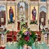 Photo of Fr. Aleksey standing in front of the entrance to the Royal Doors and facing the congregation. He's wearing a green vestment with gold trim and is reading from a prayer book. There are brightly colored flowers on the center table: magenta/white roses, yellow daisies, salmon-colored carnations, and some purple/white flowers along with a cross, an icon, and some candles. The table is covered with a green cloth.