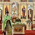 Photo of Fr. Aleksey standing at the front-left corner of the center table, with the iconstasis in the background behind him. He is facing the congregation and reading from a prayer book.