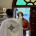 Fr. Aleksey and Joshua standing in the vestibule of the church. One of the double front doubles in the background, behind Joshua, is open. Fr. Aleksey is wearing a white vestment and has his back towards the camera. His hand is placed on top of Joshua's head while he looks slightly to the left, likely reading from a prayer book. Joshua is wearing a burnt-orange-colored shirt. There's a stained glass half-circle above the double doors, with wood between the green and brown glass pieces such that the wood pieces form what looks like the top half of a sun, with rays projected outward, and the glass pieces are between each of the rays.