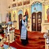 Fr. Aleksey is standing on the bottom (of three) steps leading to the Royal Doors. His left shoulder is towards the Royal Doors, and with his right hand, he is dipping a holy water brush (aspergillum) into a gold vessel containing holy in preparation to for blessing the chuch and parishioners. He's wearing a black cassock with a light blue stole (epitrakhilion) and standing on red carpet. The Sanctuary drapes and cloth on the center table are similarly blue like his stole.