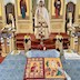 Fr. Aleksey is standing in front of the Royal Doors, facing the congregation and holding a cross in this right hand. He's wearing white vestments trimmed in gold. The center table fills the lower third of the frame, and it's covered in a light-blue cloth with several items placed on top of it, including a cross with flowers around the base, two vigil candles, and two white candles in tall, gold candle-holders. There are also two largish icons on the table in the bottom-center of the photo.