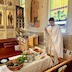 Fr. Aleksey is standing in the right half of the frame and is holding a censor that he's using to bless fruit and vegetables that are on a table in front of him. The table extends from the lower, left corner of the photo into the bottom third of the frame. There's a stained glass window behind Fr. Aleksey and a mural on the wall over his right shoulder. A pew is partially visible in the lower right corner of the photo, and part of the iconostas appears in the upper left portion of the photo. Fr. Aleksey is wearing white vestments with gold trim.