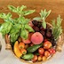 A round, wicker basket of fuit and vegetables, including peaches, cherry tomatoes, basil, plums, sweet peppers, and small, pickle-sized cucumbers. The background of the bottom half of the frame is a white, textured tablecloth, and the upper half is the back of a wooden pew.