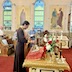 A side view of the center table with Fr. Aleksey standing in front of it, holding a prayer book while praying. His right hand is above an icon on the table, and he appears to be blessing the icon as he prays. The icon is resting on a short pedestal that is covered by a red cloth with gold trim, and there's a gold cloth on the table beneath it. There are also flowers, candles, and a cross on the table, towards the right side of the picture.