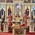Fr. Aleksey stands behind a lectern, in front of the Royal Doors. He's facing the congregation and holding up a Holy Bible. An altar server holding a candle stands on either side of him; they're wearing red and gold vestments that match his. The center table is fully visible in the foreground, at the bottom of the photo.