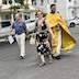 A man and a woman crossing the street with Fr. Aleksey, who is wearing a bright, golden-colored vestment. The woman is to his right, and she's wearing a black dress with large tan and white flowers. The man to her right is wearing a navy blue polo and khaki pants. Cars parked in front of houses are visible in the background.