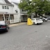 Fr. Aleksey stands with his back facing the camera as he blesses a while car in front of hime. A few other cars, spaced far apart, are visible in the photo, with houses and a couple of large trees in the background.