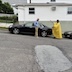 A man is standing on the side of his black car, which is to his left side as he faces the right side of the photo. The car is parked on a hill, and from the viewer's perspective, Fr. Aleksey is standing to the right of the man, on the downhill side, as he blesses the man and his car. The front of the car points uphill, to the left side of the photo, and the rectory is shown in the background. The rectory has white siding with light green bricks on the bottom.