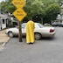 Fr. Aleksey is in the center of the frame with his back towards the camera as he blesses a tan sedan. There's a sign that says no outlet to the his left and above his head, with a sign reading watch children below it. Houses, trees, and a few other cars are visible in the background.