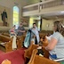 Fr. Aleksey appears in the center of frame while standing in the center aisle of the church as he blesses a young boy and a woman who are standing in the pews that appear in the lower right corner of the photo. There's also a woman holding a baby, standing in a pew across the aisle, and a man in the row behind them visible on the left-hand-side of the photo.