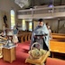 Fr. Aleksey stands at center left in the photo, holding a prayer book. There's a basket of greenery on a small table ub front of him, and another small table holding a large, silver vessel, to his right and in the lower-left of the frame. In the background are pews with parishoners.