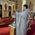 Fr. Aleksey fills the right side of the frame as he stands before a large, silver vessel in front of him wiht his hands above the vessel. The iconostas is visible in the upper-right portion of the frame, and a mural of two saints in the upper-right corner.