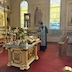 Fr. Aleksey, with his head bowed and while holding a censor in his right hand, stands center-right in the photo as he blesses the center table, which appears in the lower right-hand quarter of the photo. The table holds two icons, two gold crosses, some candles, and a plate containing holy bread.