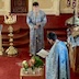 Fr. Aleksey fills most of the lower right quadrant of the photo, with his back and left side mostly shown. He holds a prayer book and is standing next to a clear, glass table that has a few vases of flowers on it along with a gold vessel for holy water. The table appears at bottom center, and there's an altar server standing such that he fills the rest of the center of the photo. He's standing far enough way from the table that his whole body is visible, and he's hold a censor. The bottom of the iconostas is visible in the background.