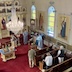 View from the choir loft showing the scene described in the photo above but from a zoomed-out perspective. Fr. Aleksey is holding a prayer book as he stands before a glass table with vases of flowers. An altar server looks on from just in front of the iconstas, and at the bottom of the three steps, while holding a censor. The center table is visible just above the lower-right-hand-corner of the photo, and several pews with parishoners fill the lower right quadrant.