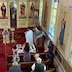 Fr. Aleksey, wearing white vestments and facing the iconostas and with his back mostly to the camera, reads from a prayer book. He's standing in front of a table covered with a white tablecloth and baskets of fruit. The table is located in front of the pews on the right-hand-side of the church, and several parishoners are seated in the pews, which fill out the bottom third of the photo.