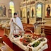 Fr. Aleksey blesses baskets of fruit on a folding table that has a white tablecloth. He's standing at center-left in the frame, with the fruit filling most of the bottom of the photo. Part of a pew appears in the lower left-hand corner, and the iconostas fills most of the photo's background.