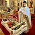 Anyone photo of Fr. Aleksey blessing the fruit. Time fills most of the left-hand-side of the photo, and he's holding a holy water vessel in his left hand and dipping a brush into the water. The fruit table appears in front of him, running diagonally across the bottom-left-corner of the frame, with the center table along the left-hand-side, and the iconostas in the background.