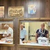Workers in the kitchen as seen through two pass-through windows in a dark-paneled wall. Only part of each window is seen, and in the left window, a man is facing left while looking down at some papers in his hand. In the right window, there's a man to the left, looking slightly towards the left (his right), and a woman on the right-hand-side smiling and looking at the camera. There are various casseroles, some bread, and cups on the counter with small tent labels describing what's in each casserole container. A few icons are hangin on the wall above the windows.