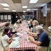 A long table, with a red and white checkered tablecloth runs through the center of the photo, from the bottom to just past halftway up the photo. There are people seated all along the table, talking and eating.