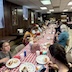 A long table, with a red and white checkered tablecloth runs diagonally from the lower left corner of the photo to about two-thirds of the way to the upper right of the photo. There are people seated all along the table, talking and eating.