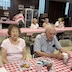 A close-up photo of a woman and a man seated at one of the tables. She's on the left and is smiling and looking at the camera, while he looks down towards the table. Two women and a man are visible at the table behind them, and they are glancing towards the camera.