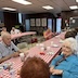 A long table, with a red and white checkered tablecloth runs diagonally from the lower left corner of the photo to about two-thirds of the way to the upper right of the photo. There are people seated all along the table, talking and eating, with a few empty chairs at the table. This is the center (of three tables), and a second table is running along behind it, in parallel, starting from the center of the left side of the frame. A few chairs from the table on the right (not pictured) are partially visible.
