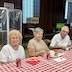 A close-up photo of two women and a man sitting at a table with a red and white checkered tablecloth. They have paused eating and are all smiling and looking at the camera.
