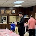 Fr. Aleksey is standing in the center of the photo, facing towards the left. He's holding a blue 5-gallon bucket with white lettering that says Lowes. He has just pulled a raffle ticket from the bucket and is reading the ticket. There's a man standing behind him, along the right edge of the frame, and his back is turned towards the camera. Part of a table with a red and white checkered tablecloth runs diagonally across the lower left corner of the photo, and dark-panel walls are in the background. Part of the kitchen can be seen through two pass-though windows in the back wall.
