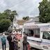 Several people are standing outside, waiting in line for ice. They appear in the lower left quadrant, and most are smiling and looking at the camera. One man has his hands raised in a pose. The white ice cream truck fills the lower right quadrant. It had red and blue embellishments - some red stripes and much wider blue stripes that have white stars along the stipes. The words Soft Ice Cream appear in red on the truck. New the passenger door, more writing say Ice Cold Soda, and Cones, with contact information on the door. Houses, trees, and a cloudy sky fill the upper half of the photo.