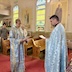 Justin Scott, the new Parish Council VP, and Fr. Aleksey are standing, facing each other and holding papers. They're both wearing light blue vestments. Parishoners can be seen standing in pews in the background, along with murals of saints on the wall between stained glass windows, and a portion of the choir loft appears in the upper left corner.