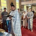 View from the front of the church, with Fr. Aleksey in the center of the left half of the frame. He's holding a prayer book. Several people are gathered around and behind him with Justin Scott standing directly behind him at a distance of about 5 or 6 feet. The others are standing closer to Justin than to Fr. Aleksey. People in pews appear in the background, and in the very back, towards the left center of frame, light is entering through main entrance to the church, where the door on the left is open.